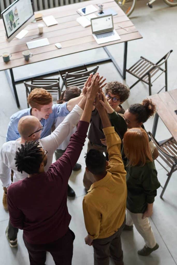 Un groupe de personnes en cercle lève les mains pour faire un geste d'équipe dans un bureau avec des bureaux et des ordinateurs portables, célébrant la collaboration et vous invitant à nous rejoindre.
