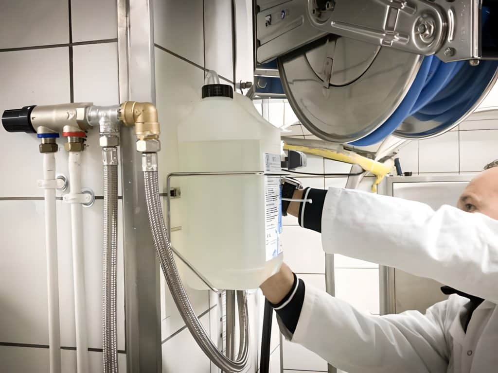 Une personne en blouse installe ou ajuste un récipient de liquide relié à des tuyaux et des tubes dans un environnement industriel carrelé, démontrant ainsi la précision et l'expertise de nos services.