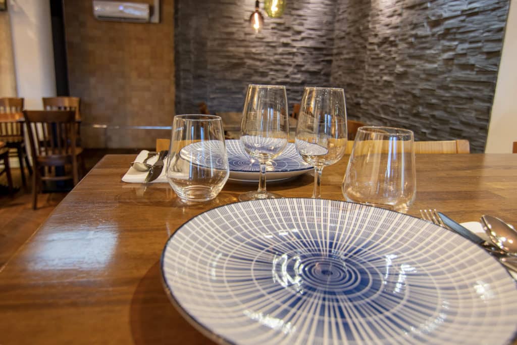 Une table à manger en bois garnie d'assiettes, de verres et de couverts à motifs, avec des chaises vides et un mur sombre texturé en arrière-plan.