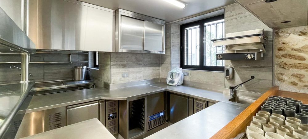 Cuisine commerciale moderne de l'espace cuisinistes, avec des surfaces en acier inoxydable, des armoires et des appareils élégants, une fenêtre avec des barreaux et un comptoir avec des tasses soigneusement empilées au premier plan.