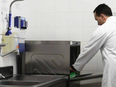 Un homme en blouse blanche place un plateau dans une machine en acier inoxydable dans une pièce propre et carrelée qui reflète la précision de nos services, avec un évier et divers équipements au mur.