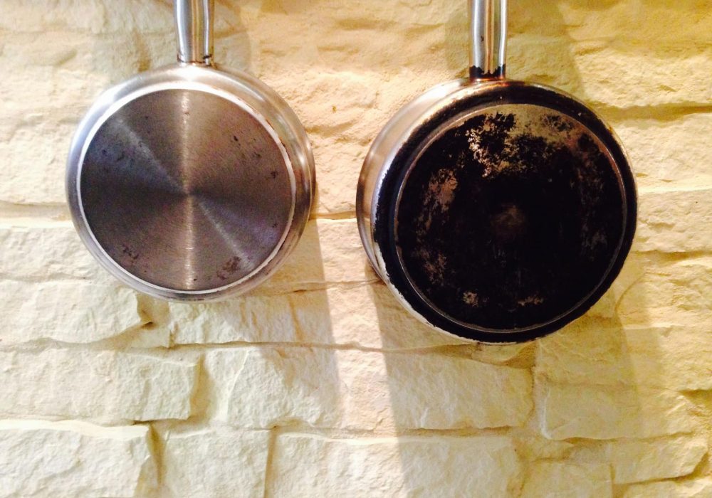 Deux casseroles en métal sont suspendues côte à côte sur un mur en pierre claire texturée ; la casserole de gauche est propre, tandis que celle de droite, marquée par la décoloration, porte les riches traces d'innombrables cuissons.