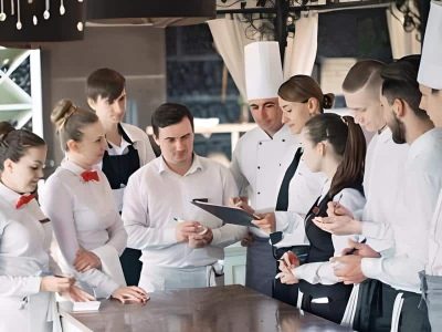 Un groupe d'employés de restaurant, comprenant des chefs et des serveurs, se tient autour d'un comptoir tandis qu'une femme munie d'un presse-papiers explique nos services et donne des instructions.