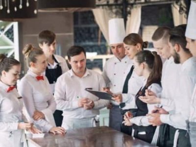 Un groupe d'employés de restaurant en uniforme se tient autour d'un comptoir, écoutant un directeur qui tient une planchette à pince et donne des instructions sur nos services.